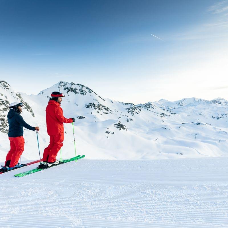 Two skiers in red gear on a snowy slope with mountain views in the Alps